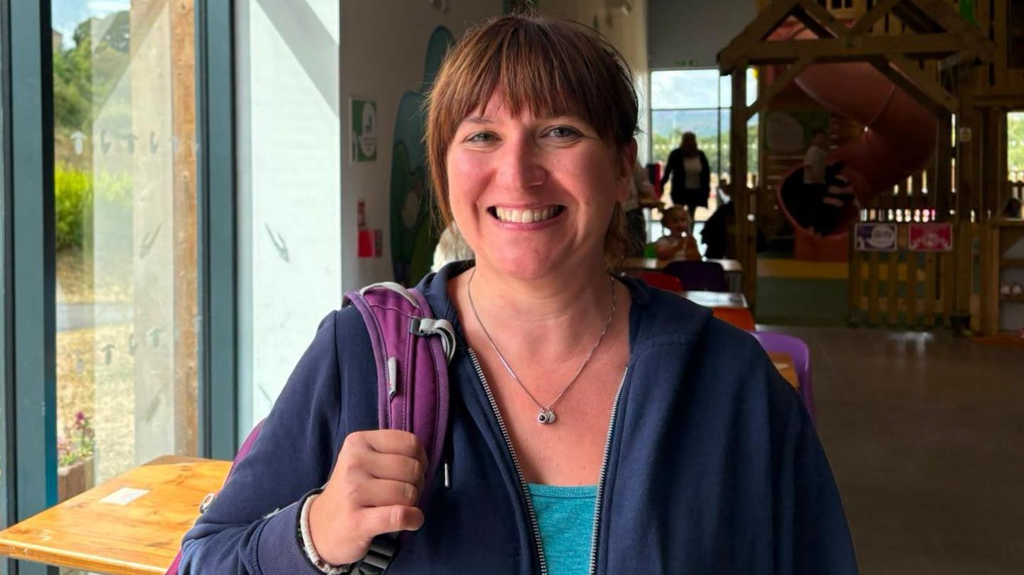 A woman wearing a rucksack, a blue unzipped hoodie and a blue top. She is stood in front of a play centre for children, slide viewable in the background.