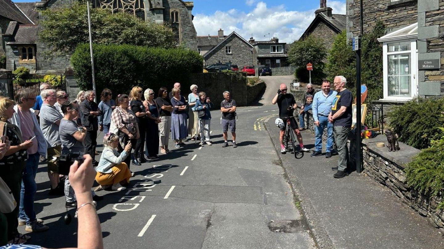 Over a dozen people standing on a road on a sunny day crowding around and taking a photo of several men next to a cat statue