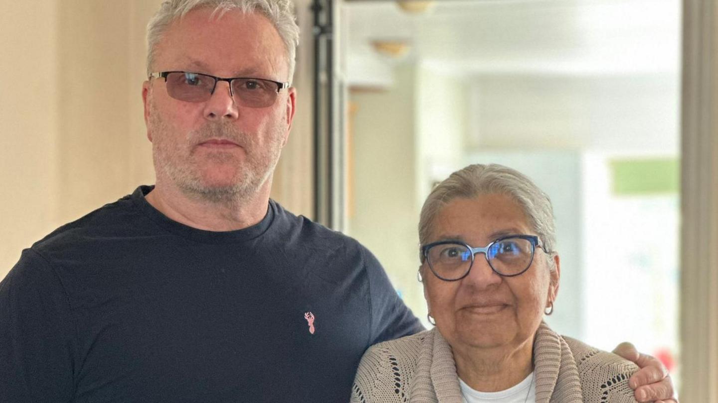 Jeff Dawes is seen in a dark t-shirt standing with his arm round Coleen, who is wearing glasses and a beige cardigan, both posing indoors and looking at the camera.