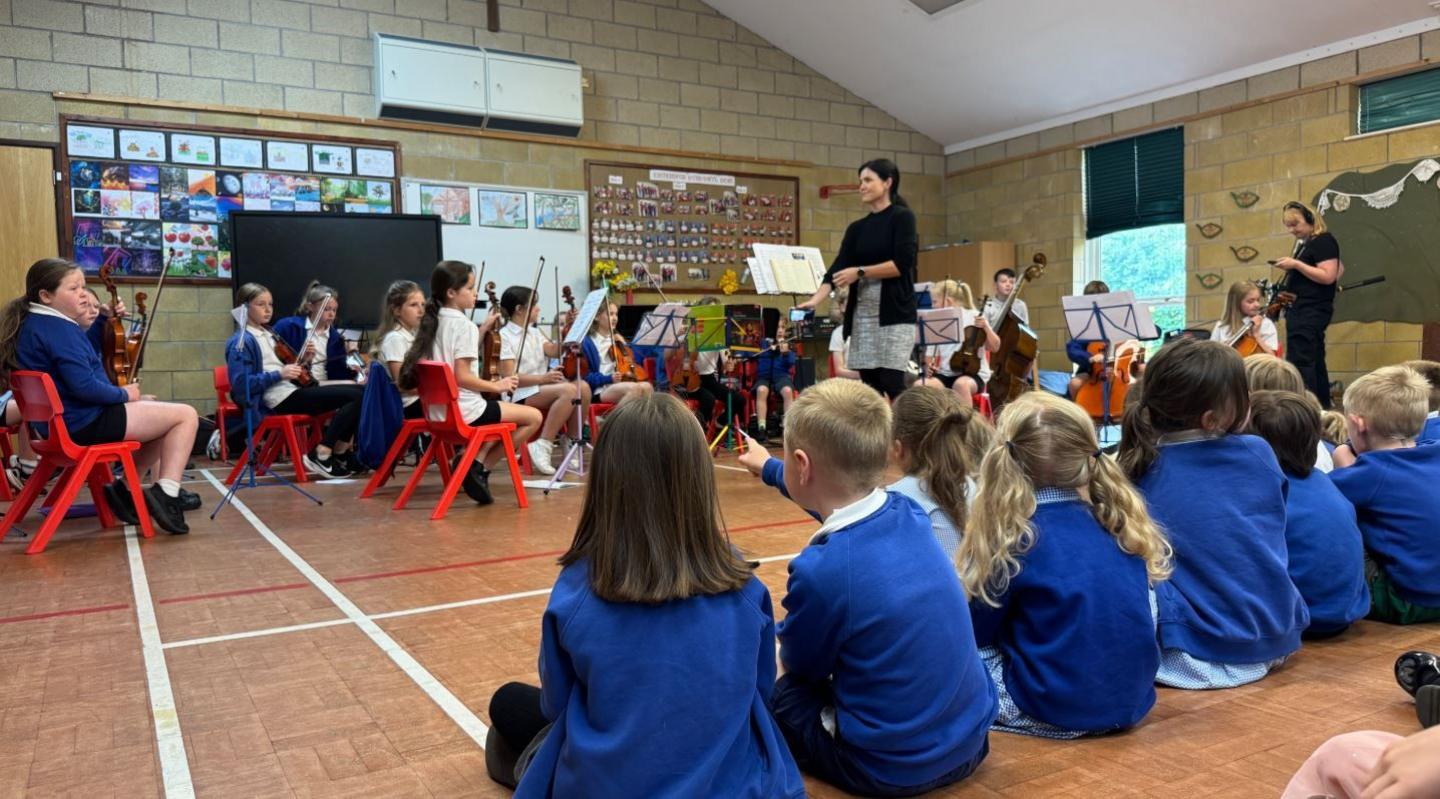 School pupils holding instruments sit on plastic chairs in a school hall, with music stands in front of them. A row of pupils wearing blue school jumpers sit on the floor watching them.