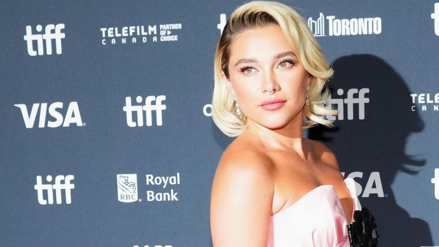 A red carpet image of Florence Pugh. She is wearing a sleeveless pink dress with a black bow at the bust.