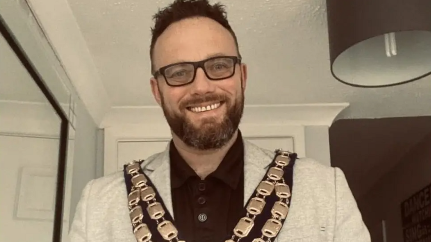 A man with glasses, dark hair and a beard in a white suit wearing ceremonial chains of office in a room with white walls