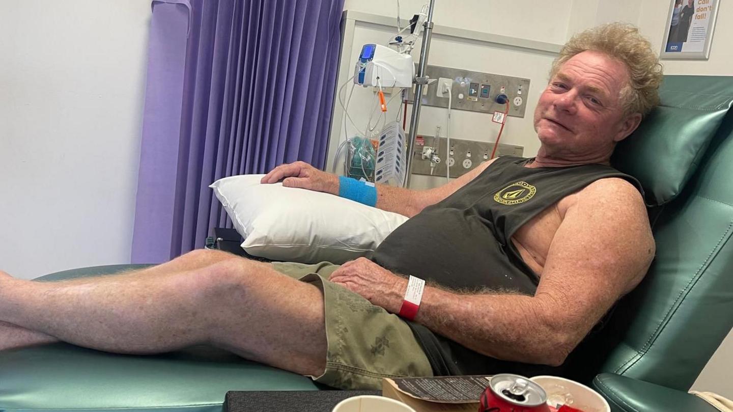 Alistair on a green hospital bed receiving chemotherapy. He is wearing a green vest and green shorts.