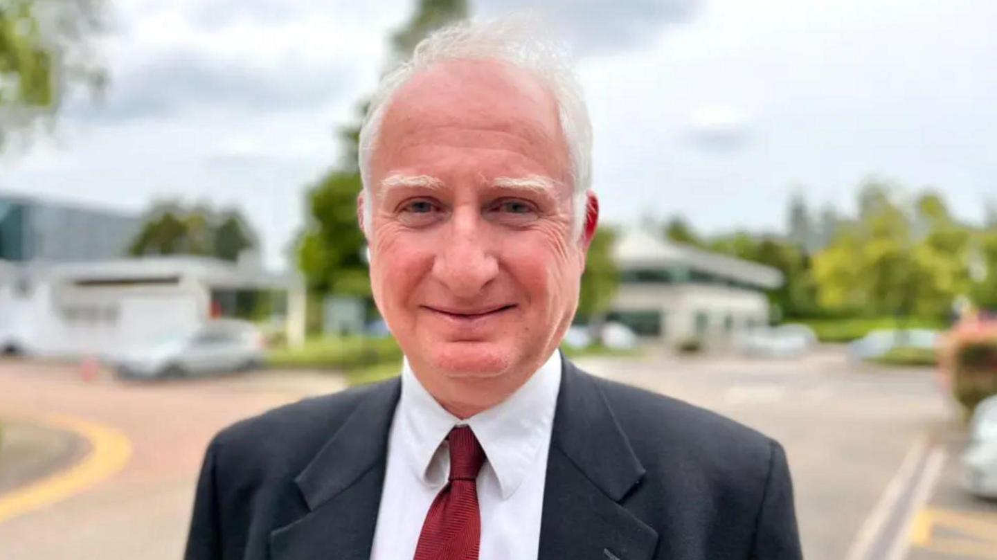 A man with white hair, a dark suit, white shirt and red tie standing in a car park