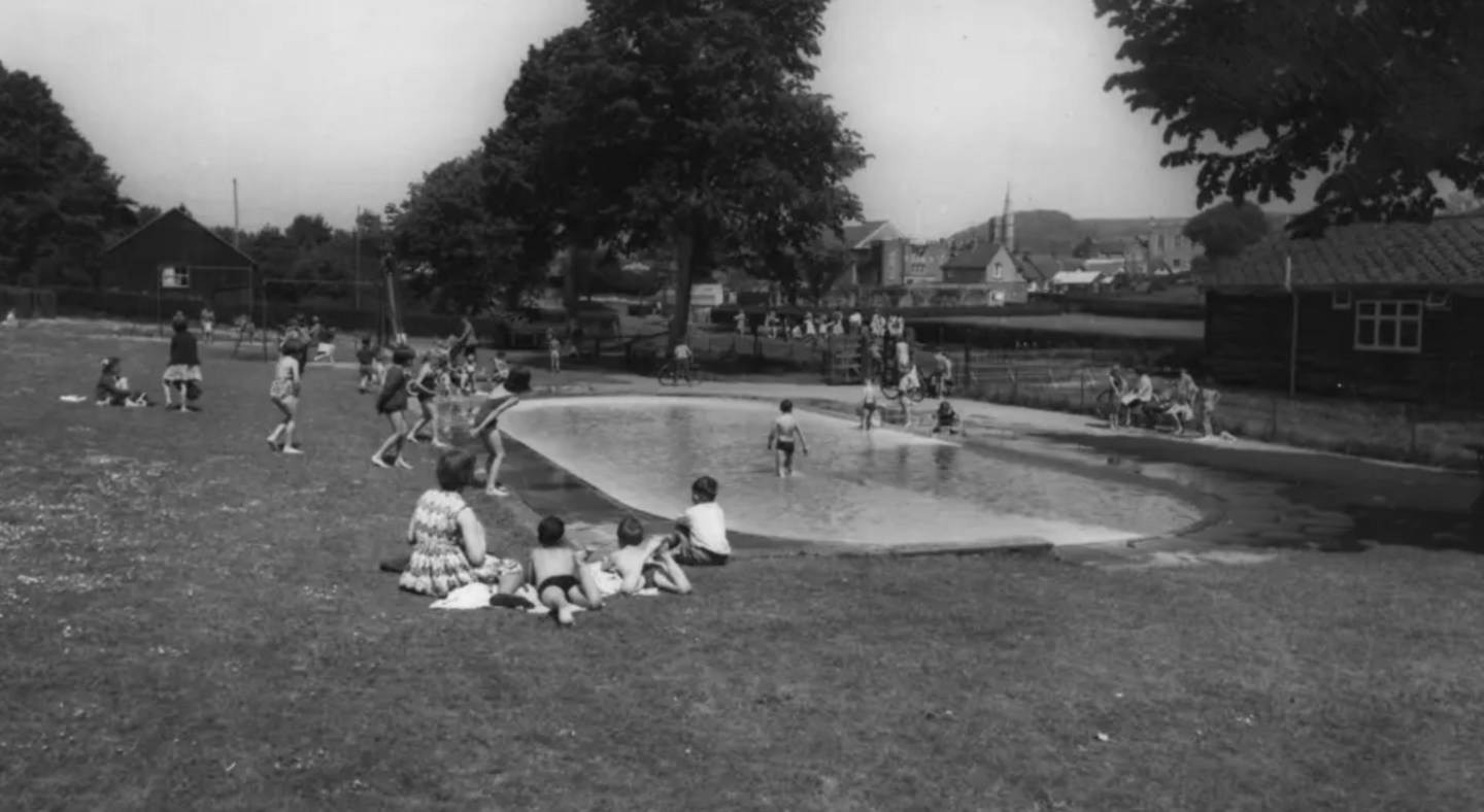 Black and white paddling pool at the Lake Pleasure Grounds from 1960, children and families sit on grass near the paddling pool.
