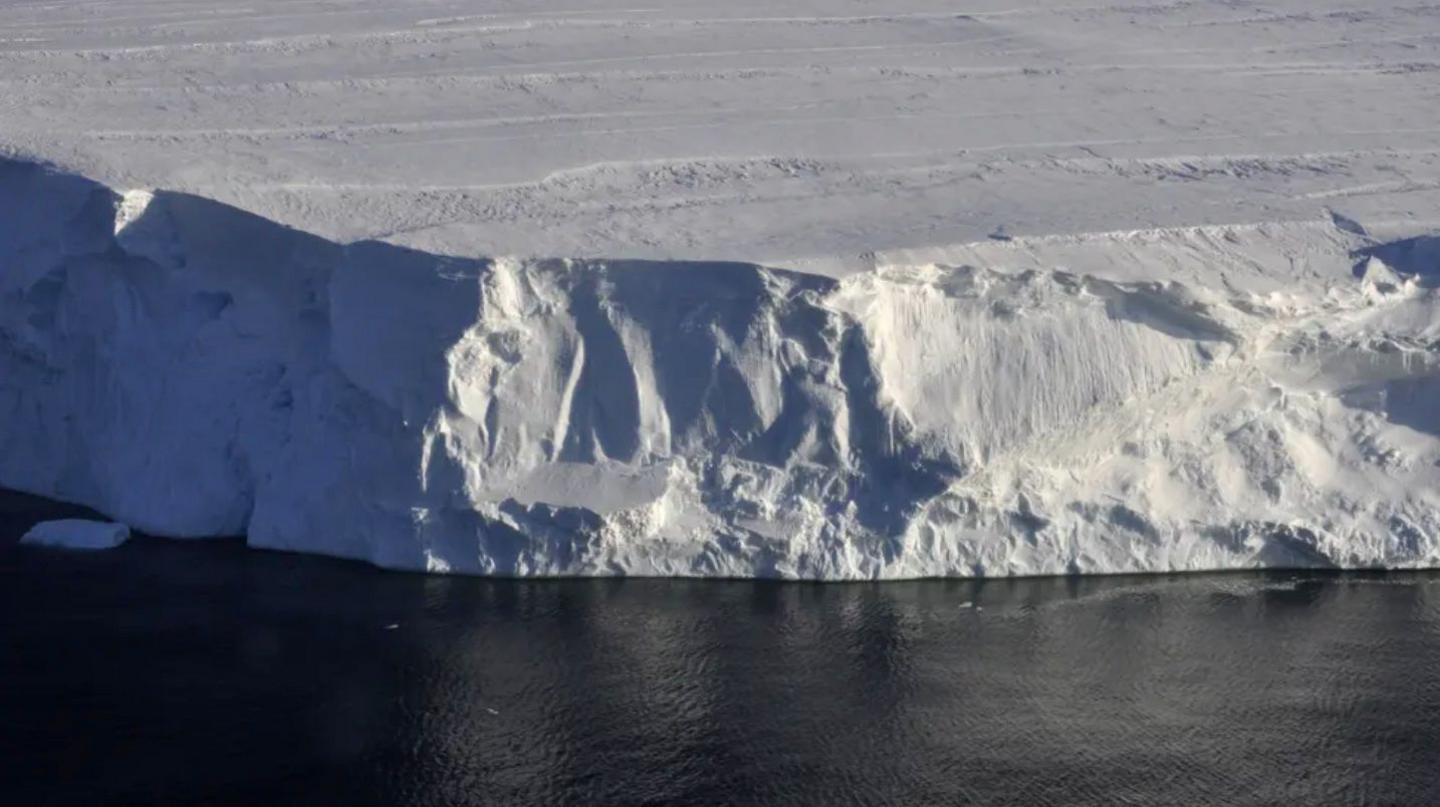 View of the front of Thwaites Glacier which appears as a white ice cliff with the dark blue ocean in front, and ice extending into the distance behind.