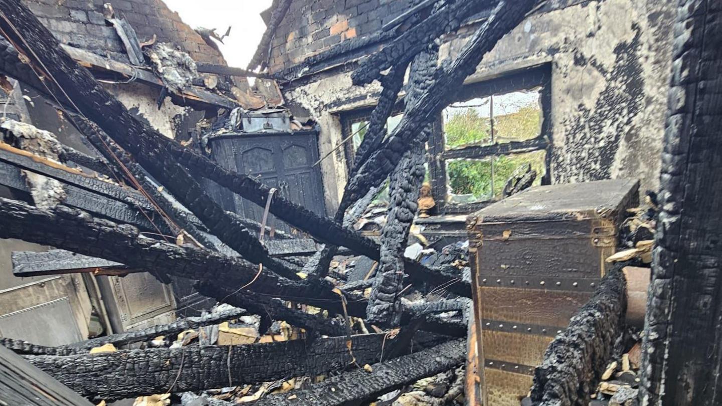 The remains of a room which is exposed to the sky following fire damage with blackened and fire-damaged beams laying on the floor and damaged items of furniture