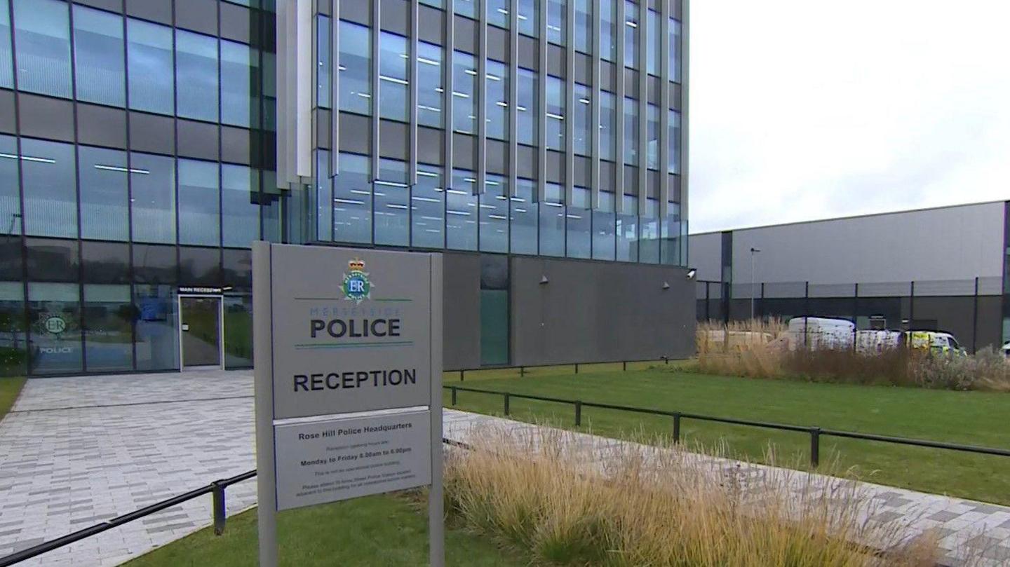 An outside view of Merseyside Police HQ on Rose Hill, which is a large rectangular office block with grey cladding around its windows and grassy areas intersected by paved pathways in front of its main entrance