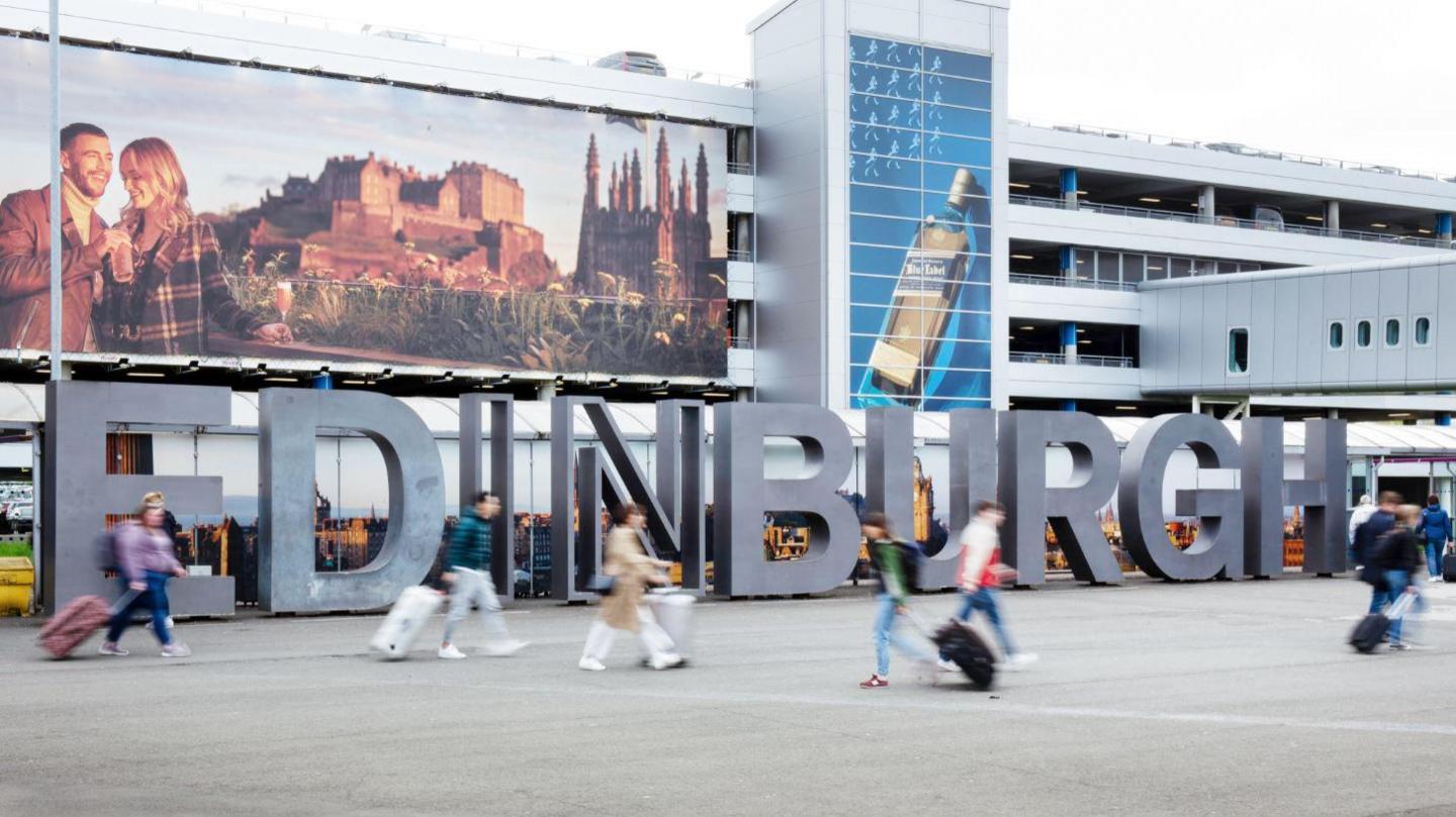 Exterior image of Edinburgh Airport, featuring a large Hollywood-style sign with "EDINBURGH" in grey block capital letters. A number of blurred passengers can be seen walking across the concourse outside the terminal building.