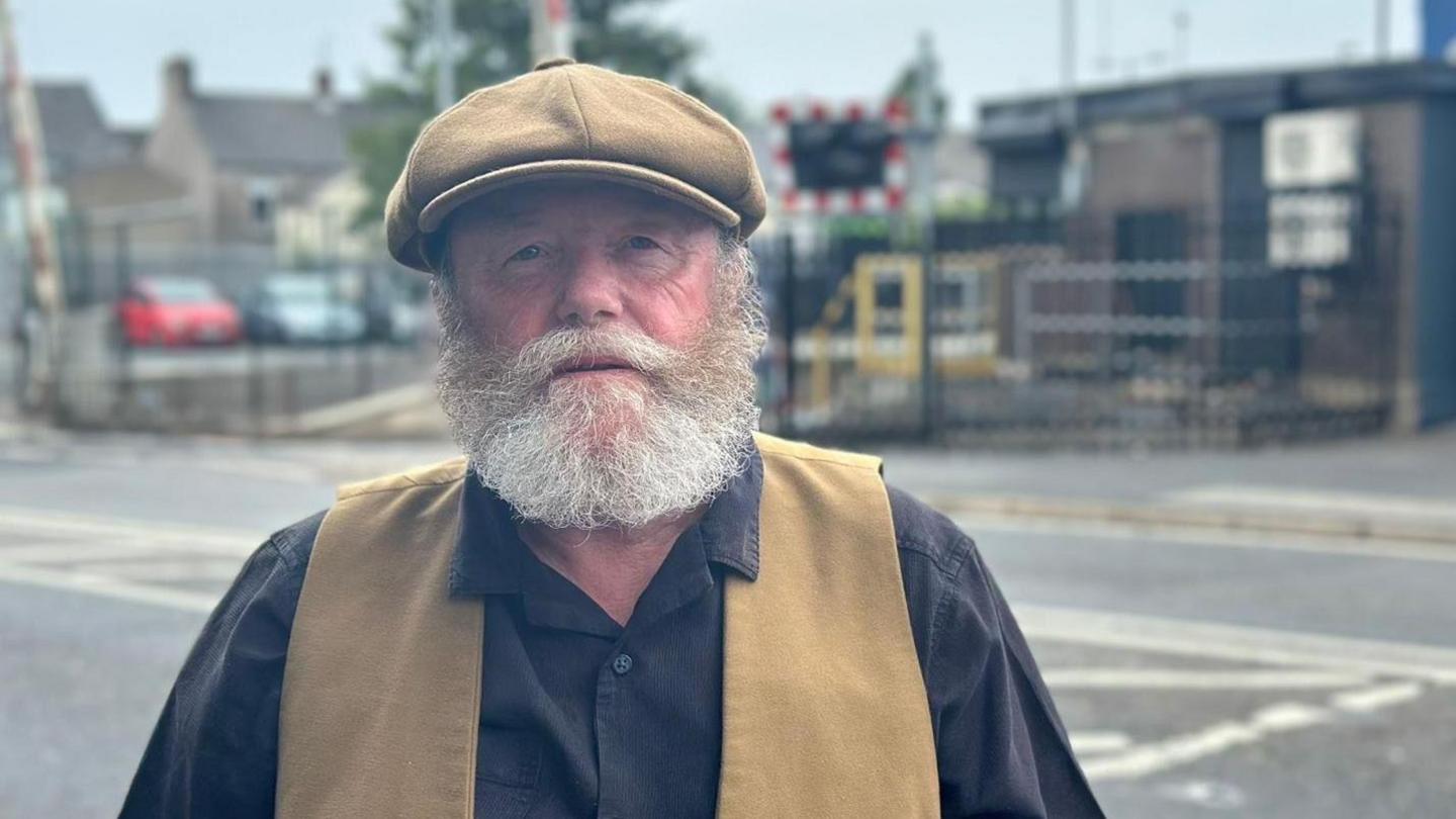 A man wears a beige flat-cap, black shirt and waistcoat of the same colour as the cap. He has a thick short grey beard. The man is standing beside a black tarmac two-lane road. Behind the road, out-of-focus, is what appears to be a train station carpark as well as the train tracks.