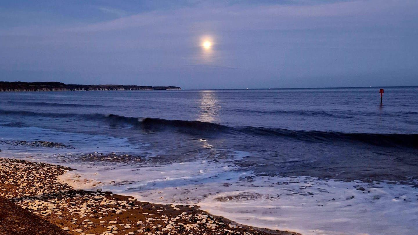 A photo of the full moon above the waves on the beach, with chalk cliffs in the distance.