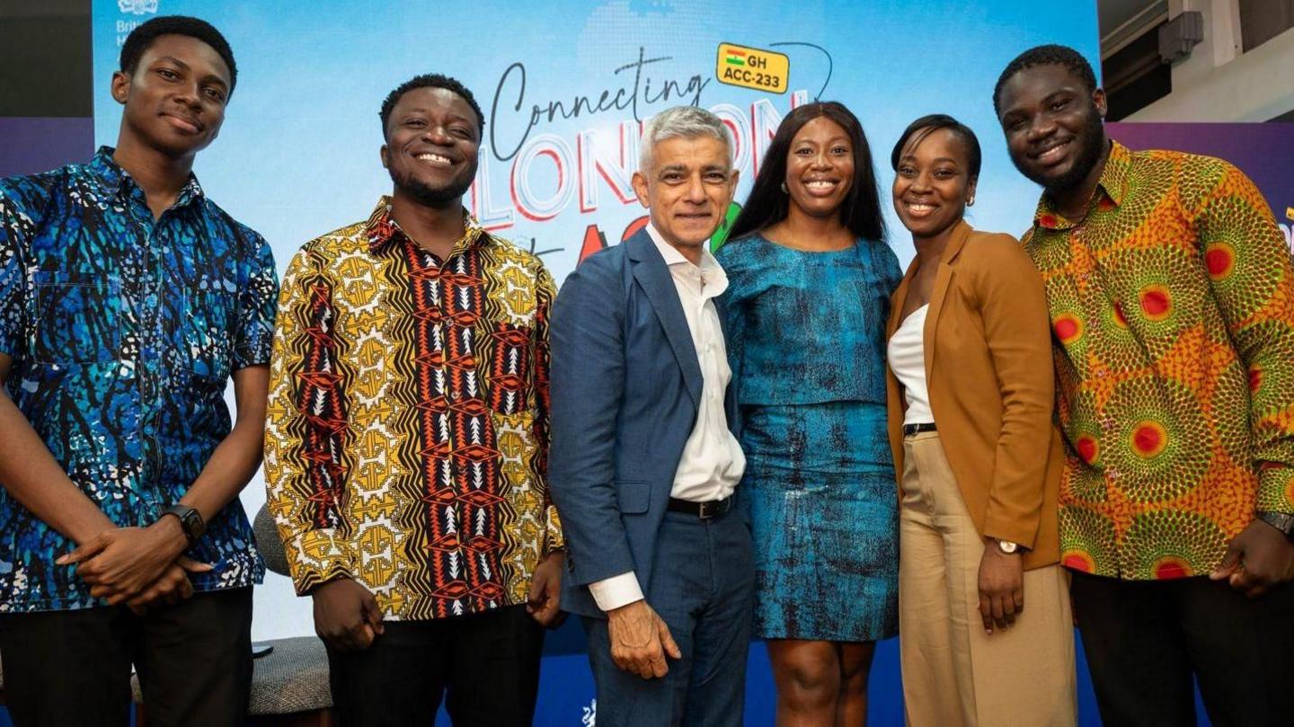 Sadiq Khan stands at the centre of a group of people posing for a photo. He is wearing a blue suit and white shirt. 