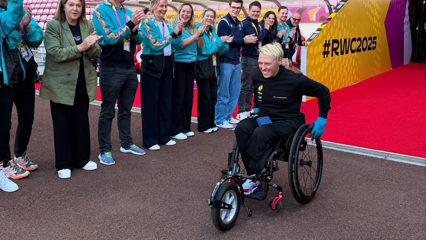 A female wheelchair user with short, bleach blonde hair pulls into a sports stadium, with a line of people clapping her to one side.