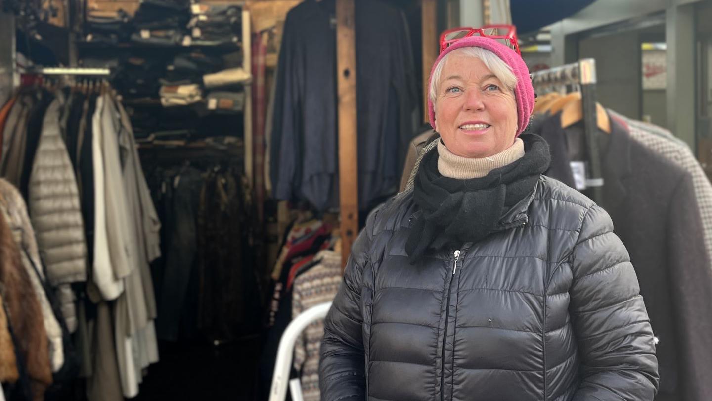 Heather Lovering wearing a black coat and pink hat. She's smiling and stood in front of her market stall.