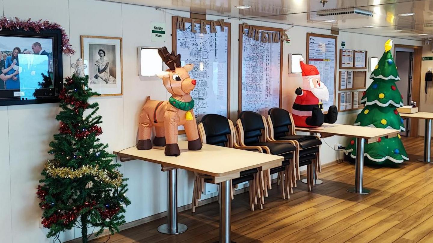 Decorations including Christmas trees and an inflatable reindeer and Santa are seen in part of a chip. There is a Merry Christmas banner on the wall, which is adorned with photographs including one of Queen Elizabeth II