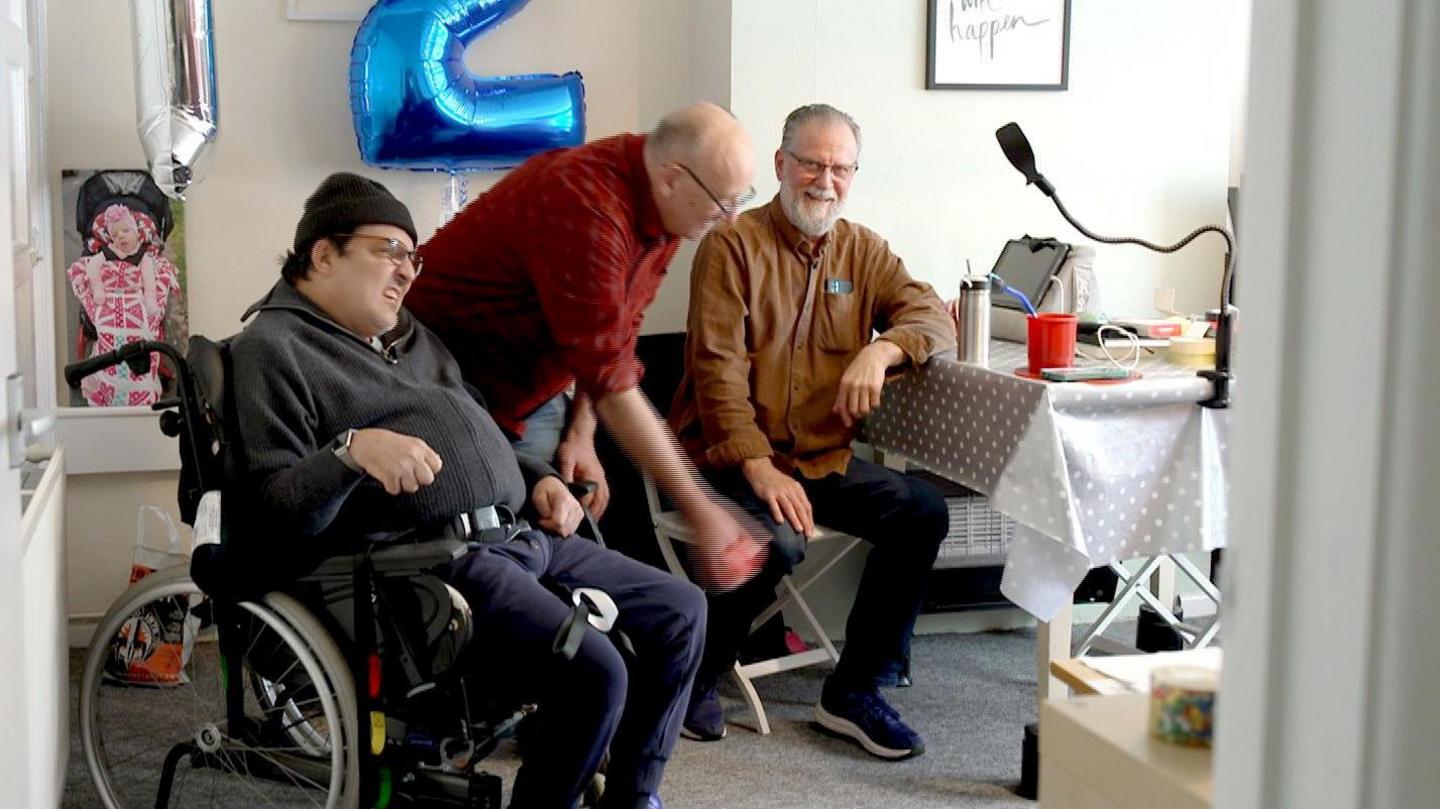 Three men, one of them in a wheelchair, smile as they sit in a room next to a table with a white tablecloth over it. They are playing a game that involves throwing an object a small distance.