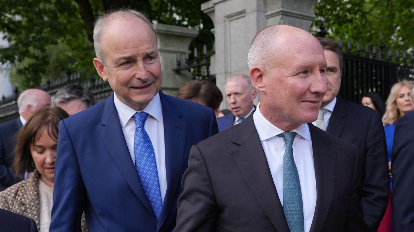 A bald Micheál Martin in a blue suit, white shirt and blue tie, stood beside a bald man wearing a black suit, white shirt and tie, they are pictured in a tight media scrum outside a fence