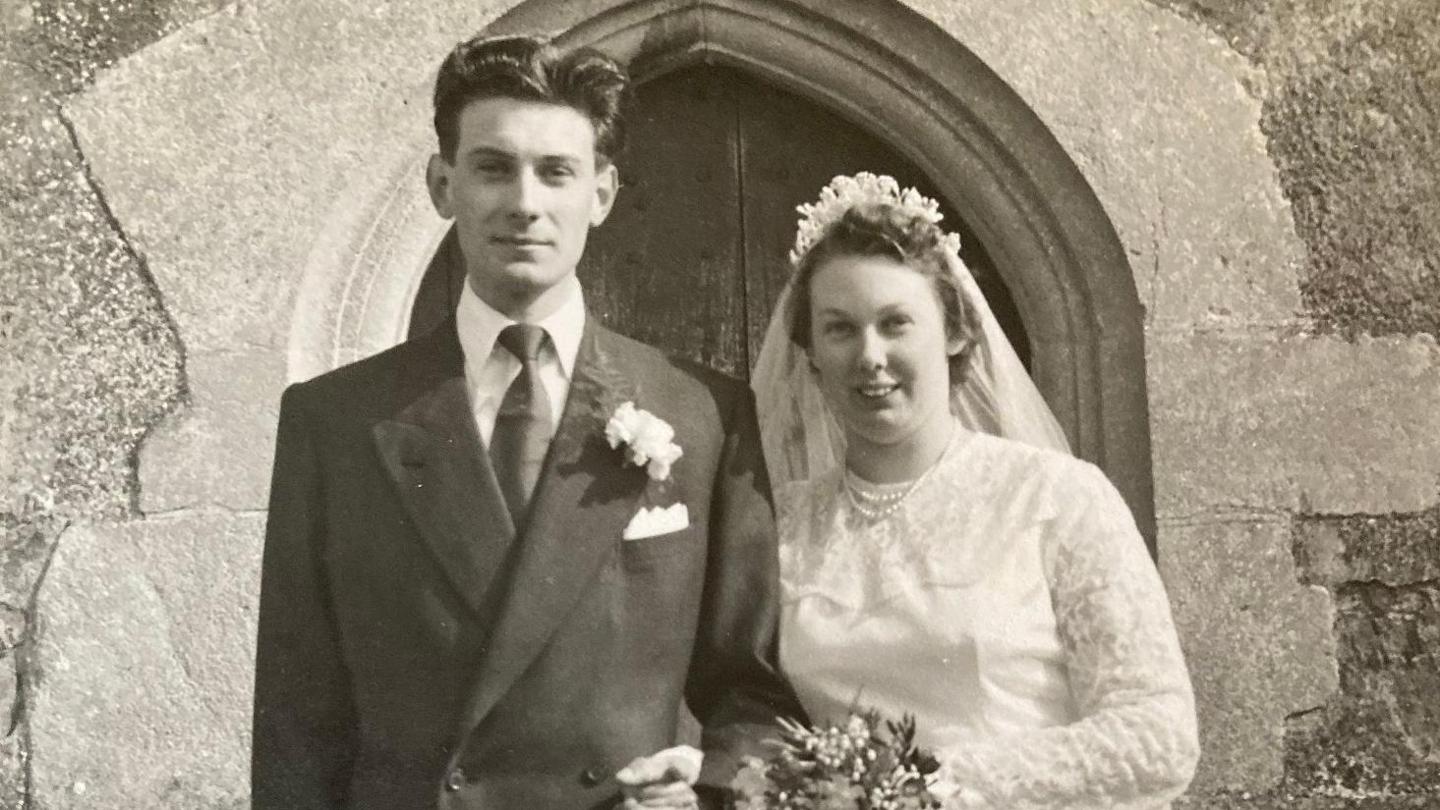 Derek and Brenda on their wedding day standing in a suit and white wedding dress with lace sleeves outside St Dunstan's Church in Baltonsborough near Glastonbury in Somerset. 