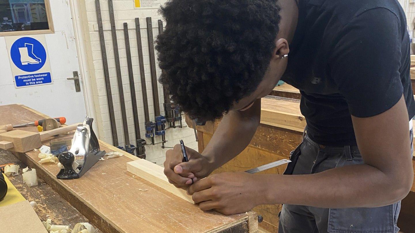 A student at Newark College measures out a plank of wood, preparing to make a cut.