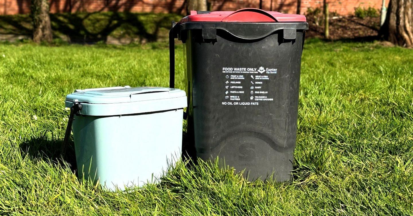 A small green caddy for food waste collection in the home and a larger black caddy with a red lid to be placed outside. Both are outside on grass.