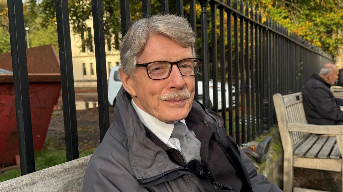 An older man with grey hair and glasses sits on a wooden bench outdoors. He wears a dark grey jacket over a black jumper and white shirt. Behind him, a black iron fence lines a path with another man sitting further along. The setting includes autumnal trees and a historic stone building with arched windows.