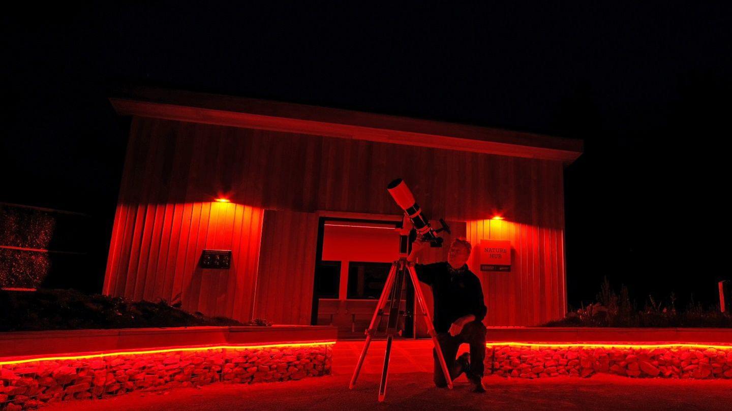 A man stars into a telescope kneeling in front of a wooden hut lit up in red lights.