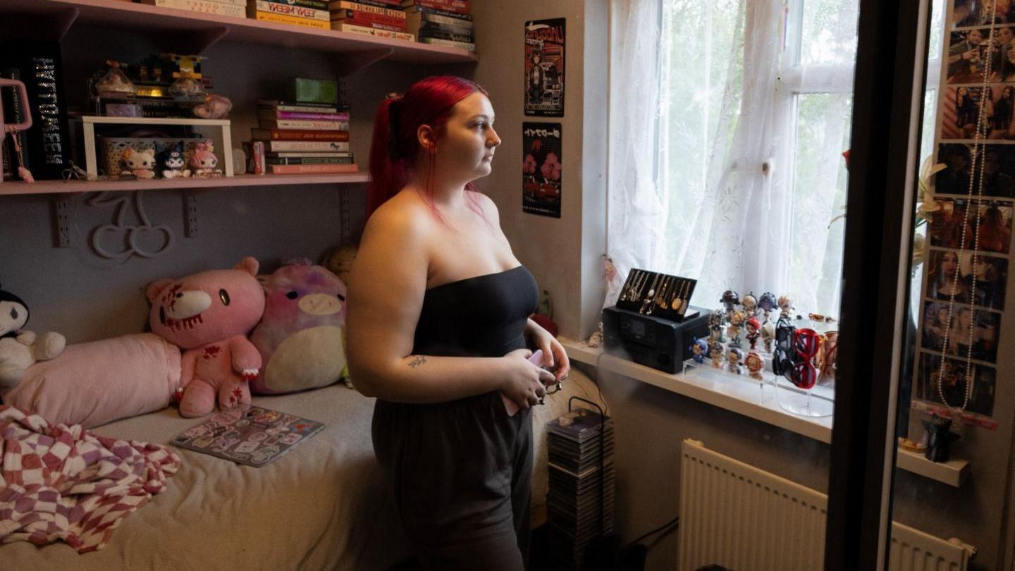 A woman with red hair standing up in her bedroom, looking out of the window, away from the camera. Her bed is behind her, covered in blankets and soft toys, with mini-figures and books on her shelves and windowsill.