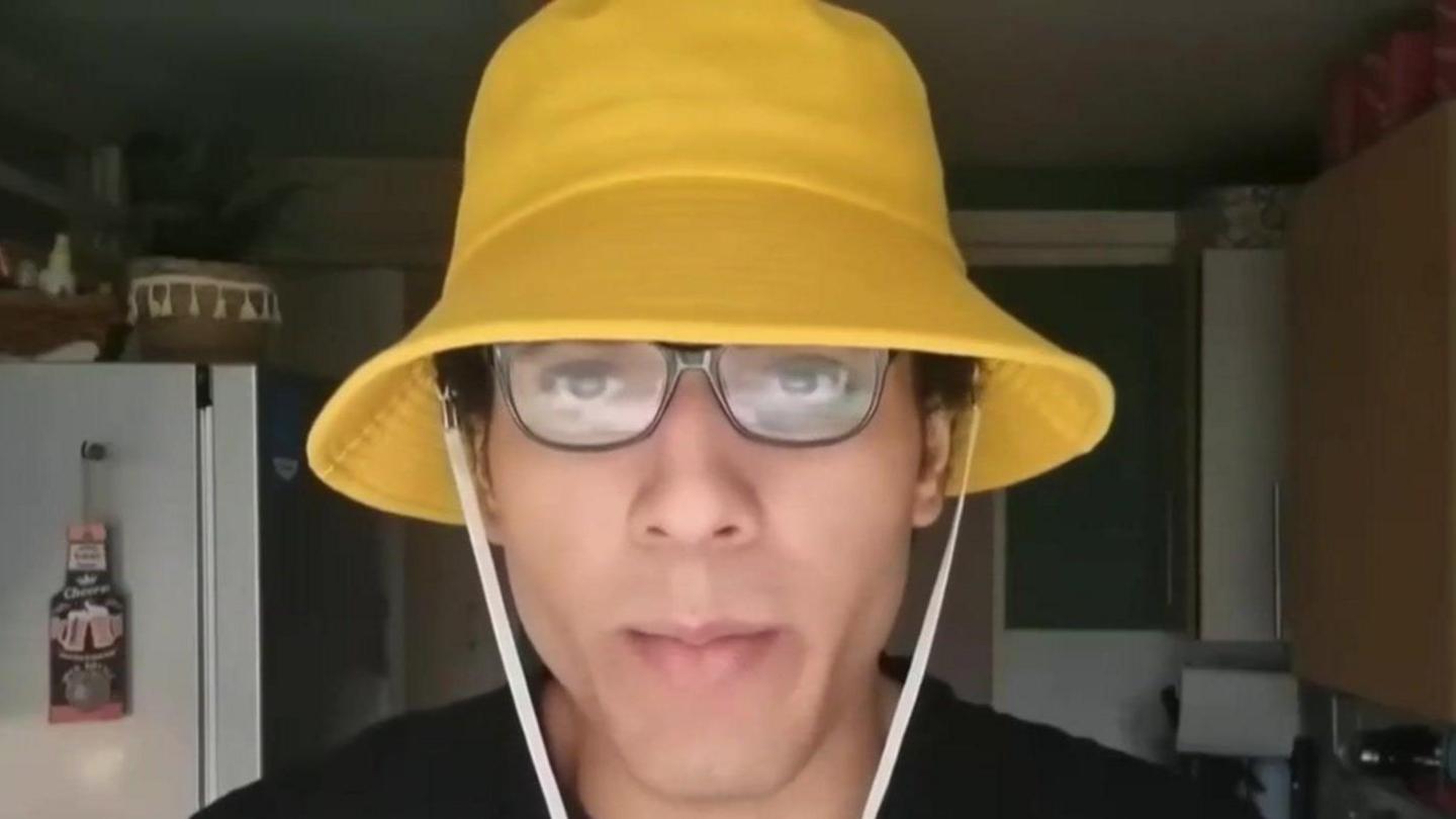 Nicholas Prosper, wearing glasses and a yellow bucket hat with a white string, talking into the camera in a homemade video in a kitchen.