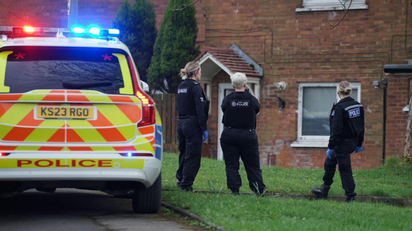 A yellow and red liveried police car with lights switched on is parked on the left. Three policewomen, in black uniform and blue gloves, stand on the lawn in front of a house