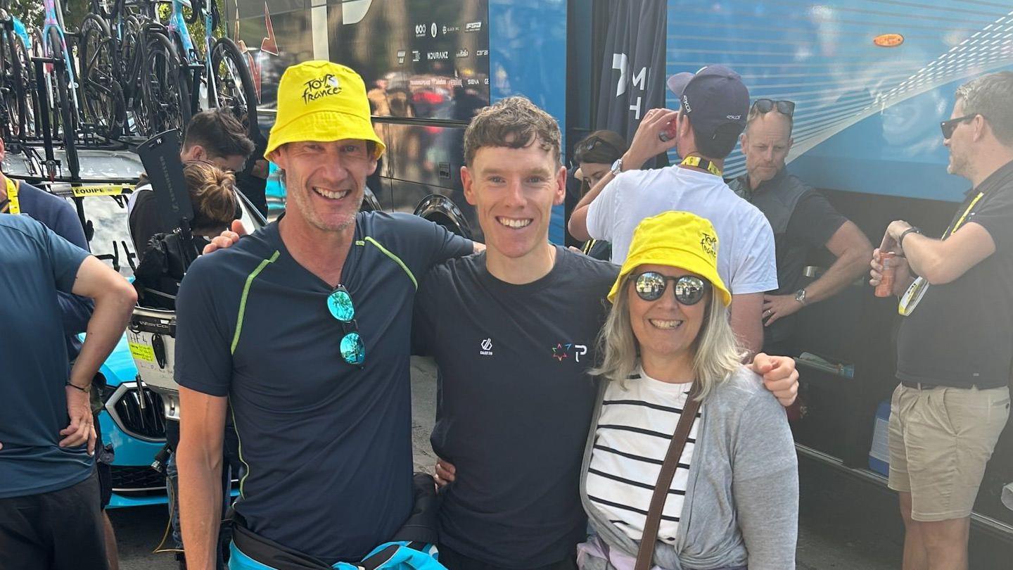 Two men in dark blue tshirts and a woman in a white and black striped shirt and grey cardigan are standing in a line with their arms around each other and are smiling at the camera. The man and the woman on each end of the row are wearing yellow bucket hats