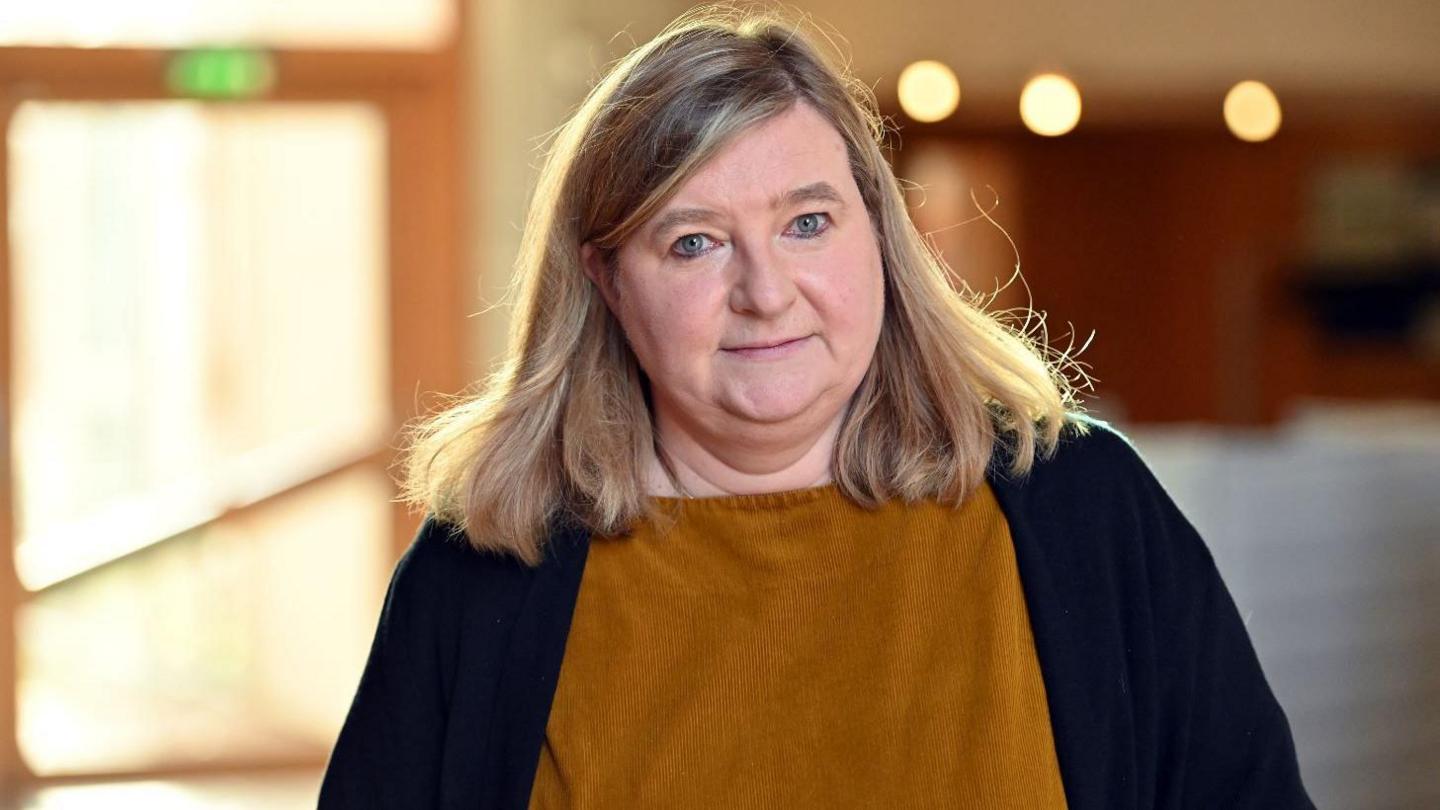 Jenny Minto, a woman with shoulder length blonde hair, looks at the camera as she walks along a corridor. She is weaing a brown top and a black cardigan
