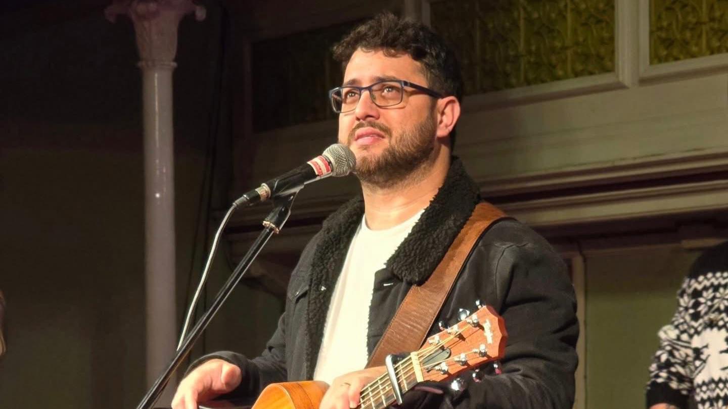 A man with short black hair and glasses is stood behind a microphone holding a guitar. He's looking out on to the audience in front of them.