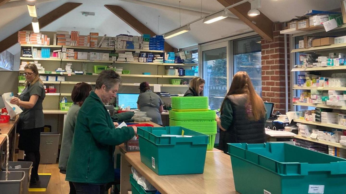 The inside of Gompels Pharmacy shows shelves of medication and a number of staff members inside, some wearing grey tops and others in green fleeces, finding medication for patients.