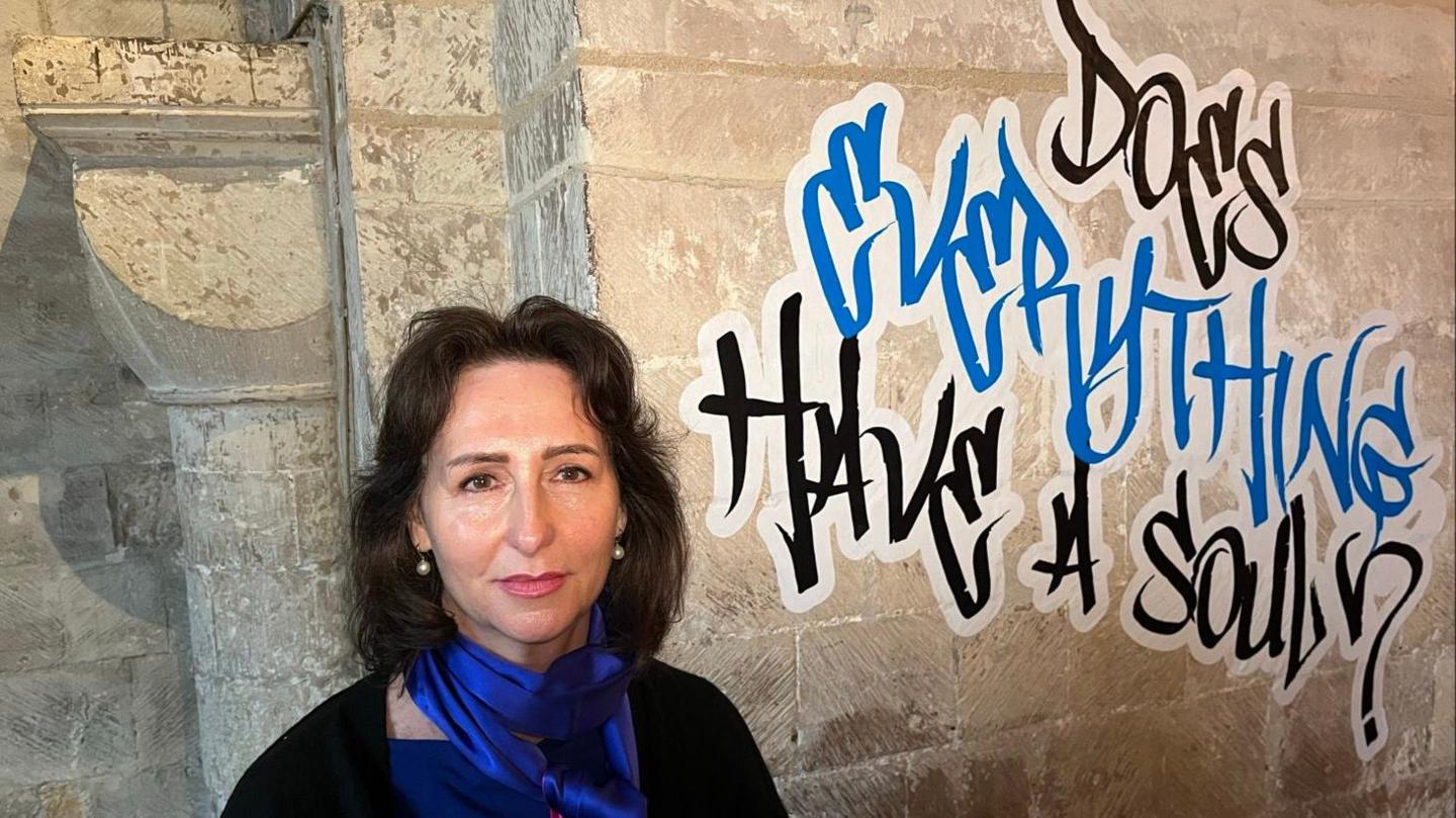 Curator Jacqueline Creswell standing in Canterbury Cathedral crypt beside a graffiti style artwork that reads 'Does everything have a soul?'
