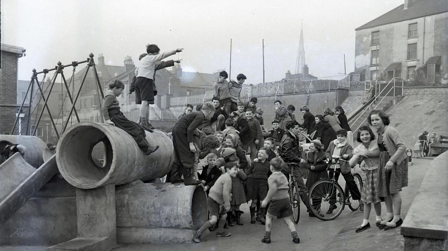 A group of children climb over play equipment in a black and white photo of a playground in Devonport. Some are holding bicycles and smiling at the camera.