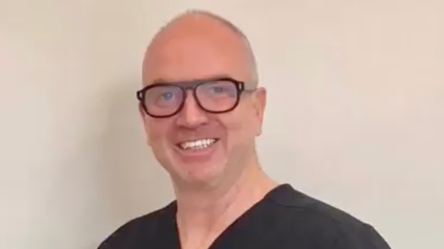 Neil Counihan is smiling into camera, in a head and shoulders photo. He's wearing black scrubs, chunky dark framed glasses and is mostly bald.