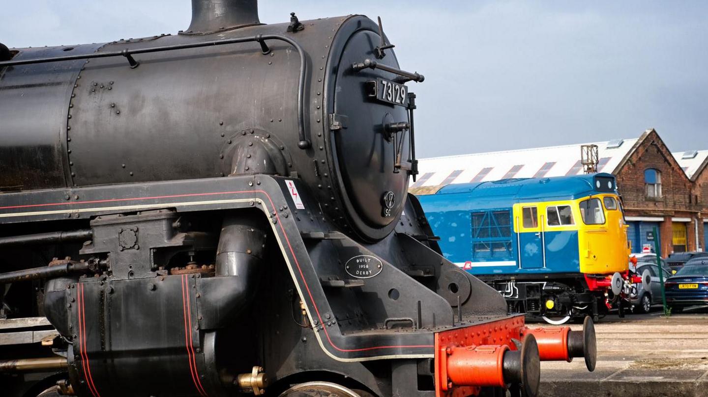 A black steam locomotive is parked in the foreground, with a blue and yellow train and brick industrial buildings visible in the background