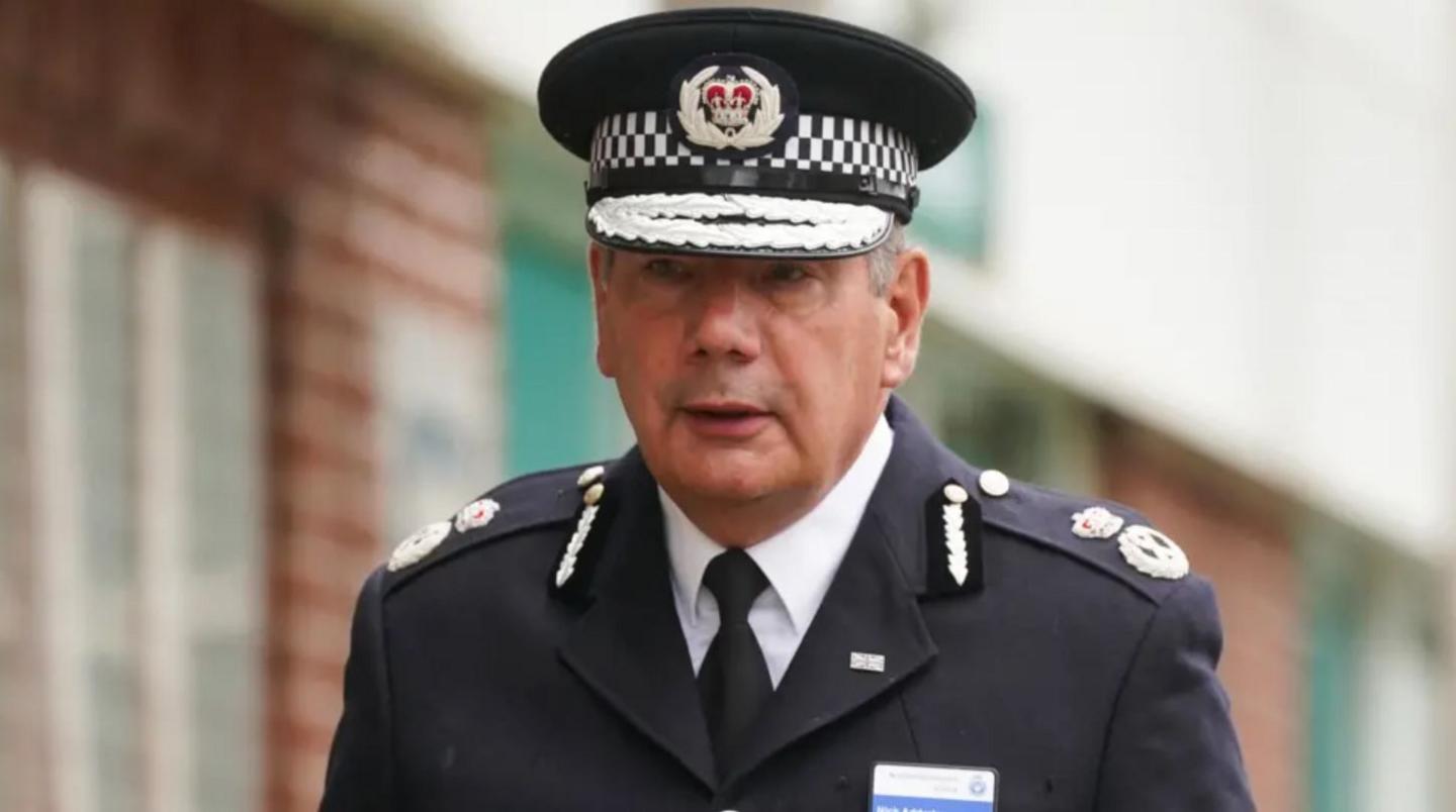 Man in police chief's uniform and cap. He is seen walking outside a building. There are epaulettes indicating his rank. The uniform is black with silver insignia.