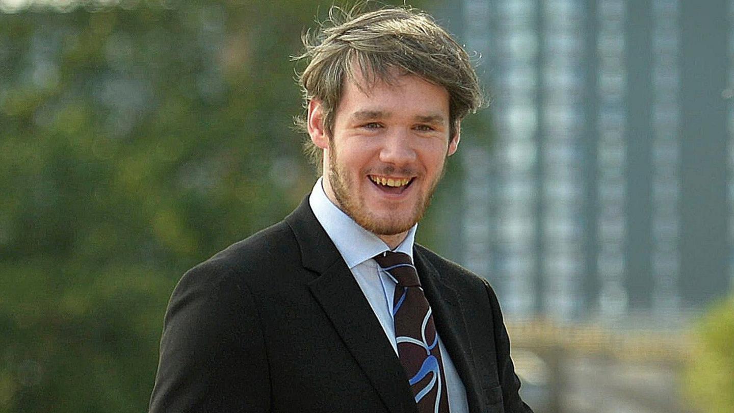 A smiling young man who has sandy hair and a beard, wearing a black suit jacket, a light blue shirt and a brown and blue patterned tie. He is outdoors, with trees and a tall building blurred in the background.
