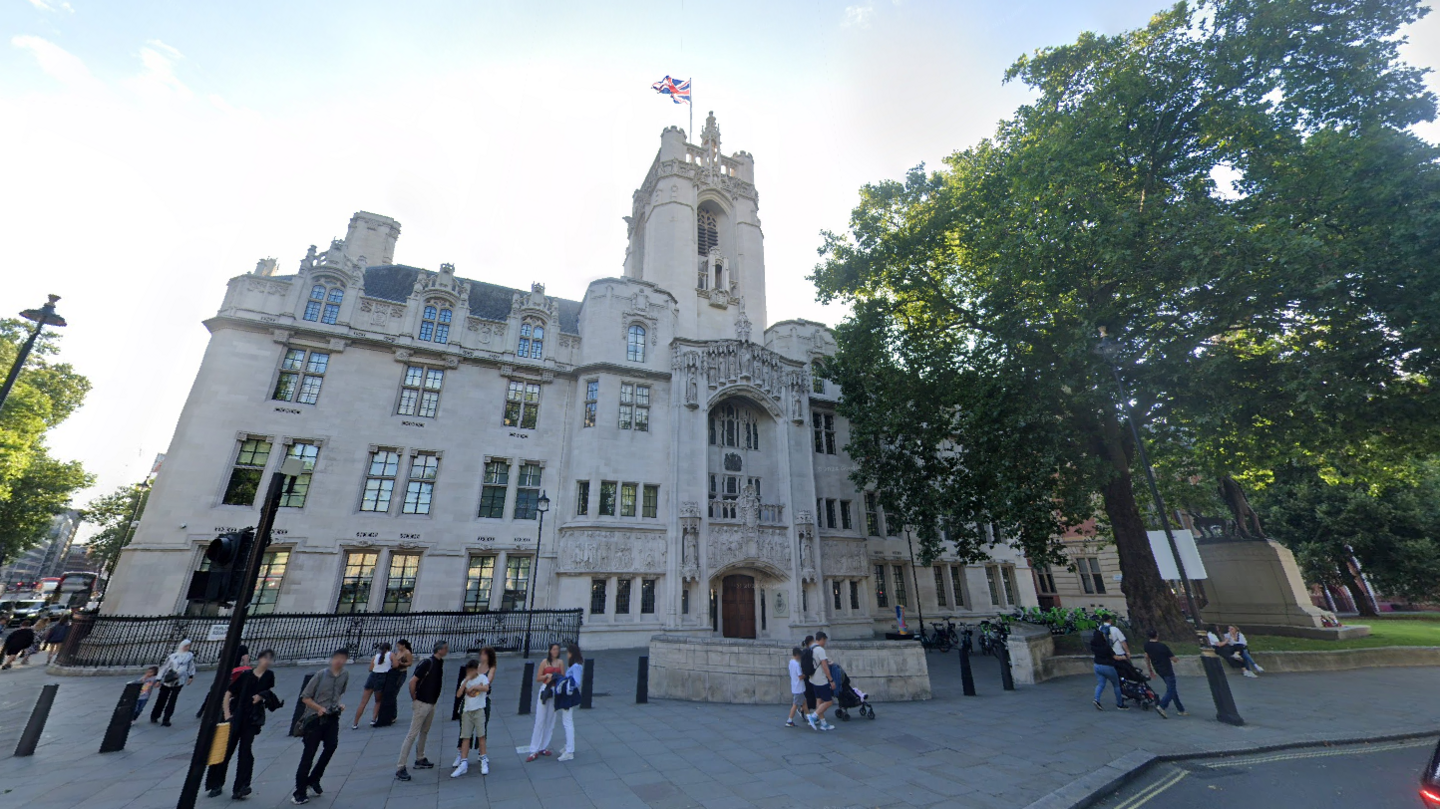 An image of the outside of the Supreme Court in London. It is a large white-coloured building with a tower and a Union flag flies from a mast on top. In front of the building there are people standing on the pavement. There are trees either side.