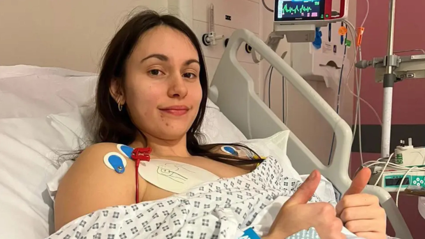 A woman with long dark hair in a hospital gown lying in a hospital bed with both thumbs up