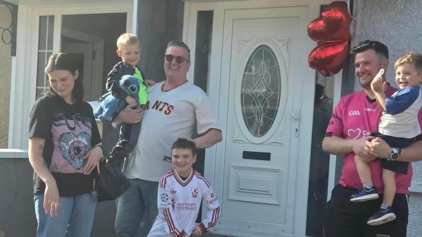 Dáithí with his family outside their home. From left to right - a woman white dark hair is wearing a black t-shirt and blue jeans and carrying a handbag, a young boy with blonde hair carrying a Stitch teddy bear is being held by a man with short brown hair wearing sunglasses, a cream Giants t-shirt and blue jeans, Dáithí has short brown hair, wearing a white long sleeve Liverpool jersey, a man with dark hair is wearing a pink GAA jersey and black trousers and carrying a young boy wearing a blue and white top, navy shorts and trainers.