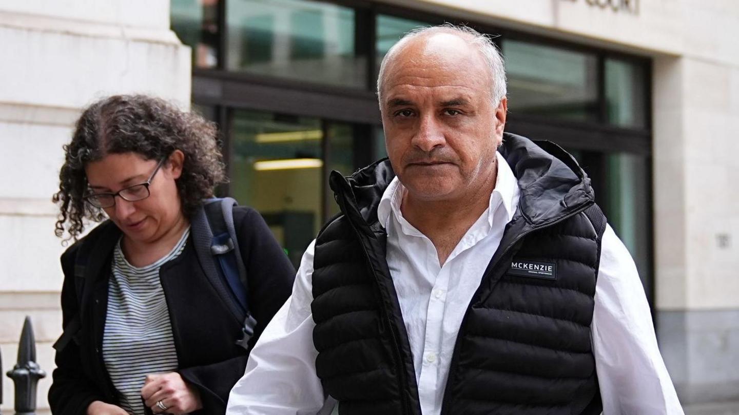 A middle aged man wearing a white shirt and a black gilet looks into the camera as he walks down the street outside a magistrates court. 