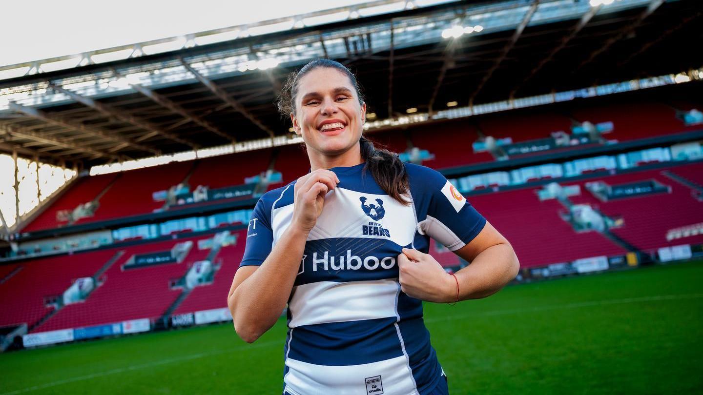 USA rugby player Ilona Maher stands on the pitch at Ashton Gate, smiling at the camera as she holds her Bristol Bears club crest on her shirt. In the background the stadium's main stand can be seen. The picture is taken on a clear day