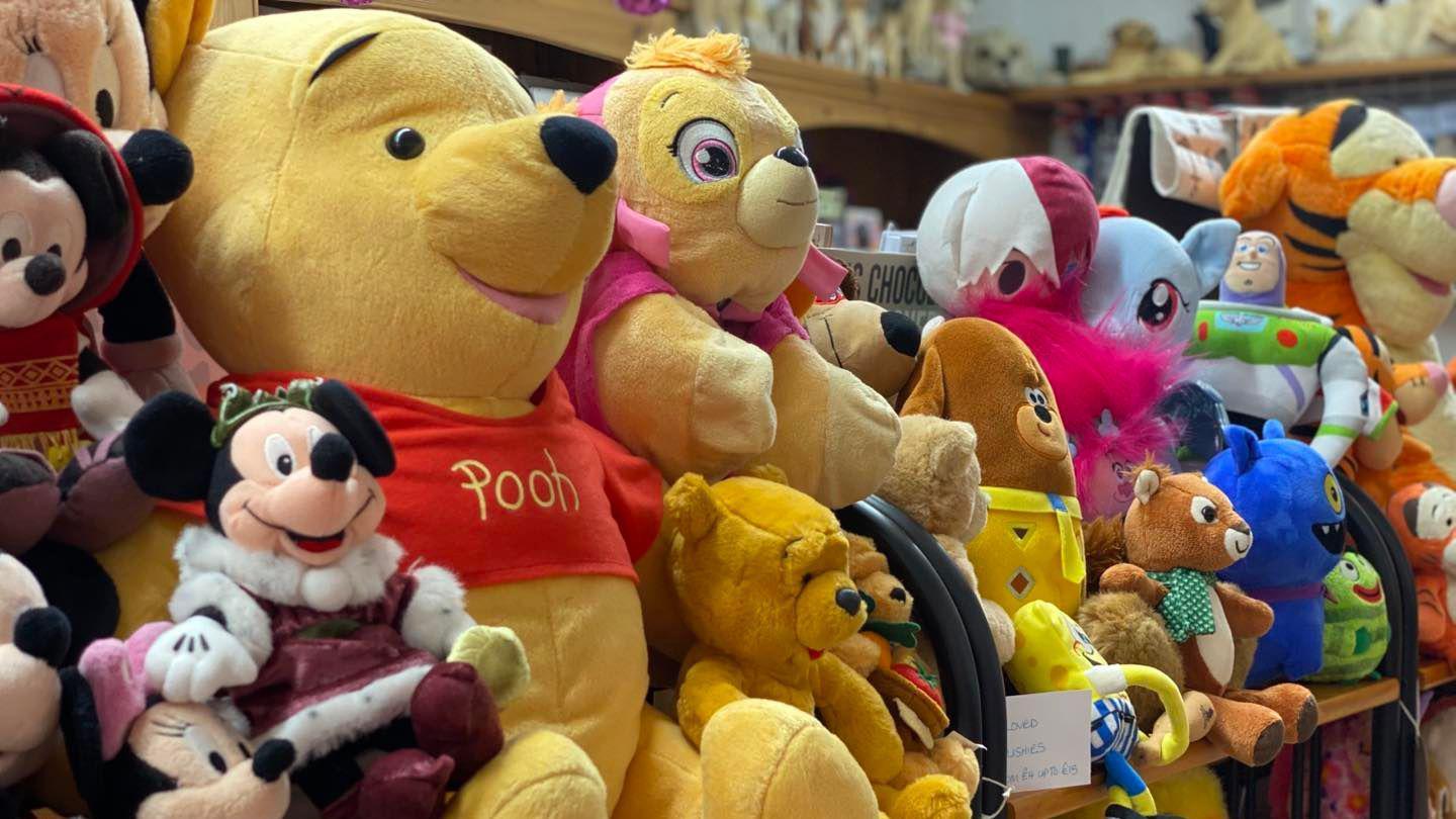 A set of soft toys lined up on a shelf in the card shop. There is a very large Winnie the Pooh on the left and a similar-size Tigger on the right. Next to Pooh are several Minnie Mouse toys. There is also a Duggee, Buzz Lightyear and Spongebob Squarepants, among others.