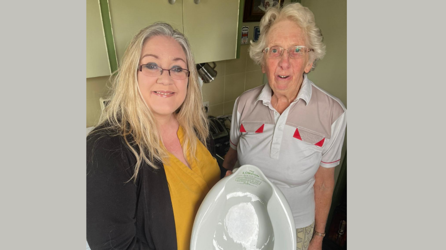 Julie Stevenson with long blonde hair and glasses stands next to Mrs Jacobs. Mrs Stevenson is wearing a yellow top with a dark green cardigan. Mrs Jacobs is wearing a white polo top with some red details on it. 