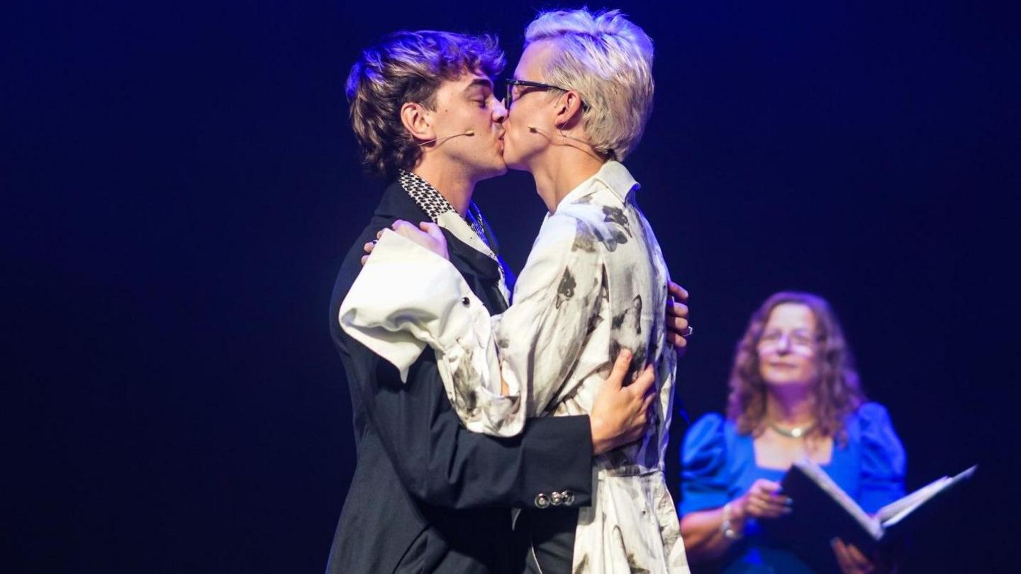 A well-lit couple kiss on stage against a dark background as a female wedding celebrant - with long brown hair, glasses and wearing a blue dress - looks on in the background. She is holding an open hard-backed book. Linus - who is wearing a flamboyant white shirt with an oversized half collar on the right hand side and dark rimmed glasses - has blonde hair combed in a quiff. Joseph is wearing a short navy jacket which has a large collar and a white, grey and black neck scarf. He has brown/ blonde hair.