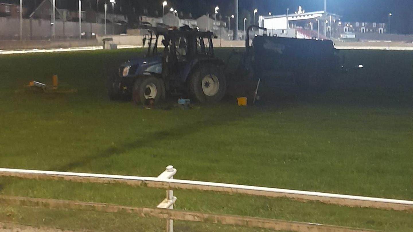 A blue tractor sits in the middle of a dog racing track in Derry. White fencing can be seen in the foreground, and a spectator's stand is visible in the background. It is night time and the stadium is flood lit.