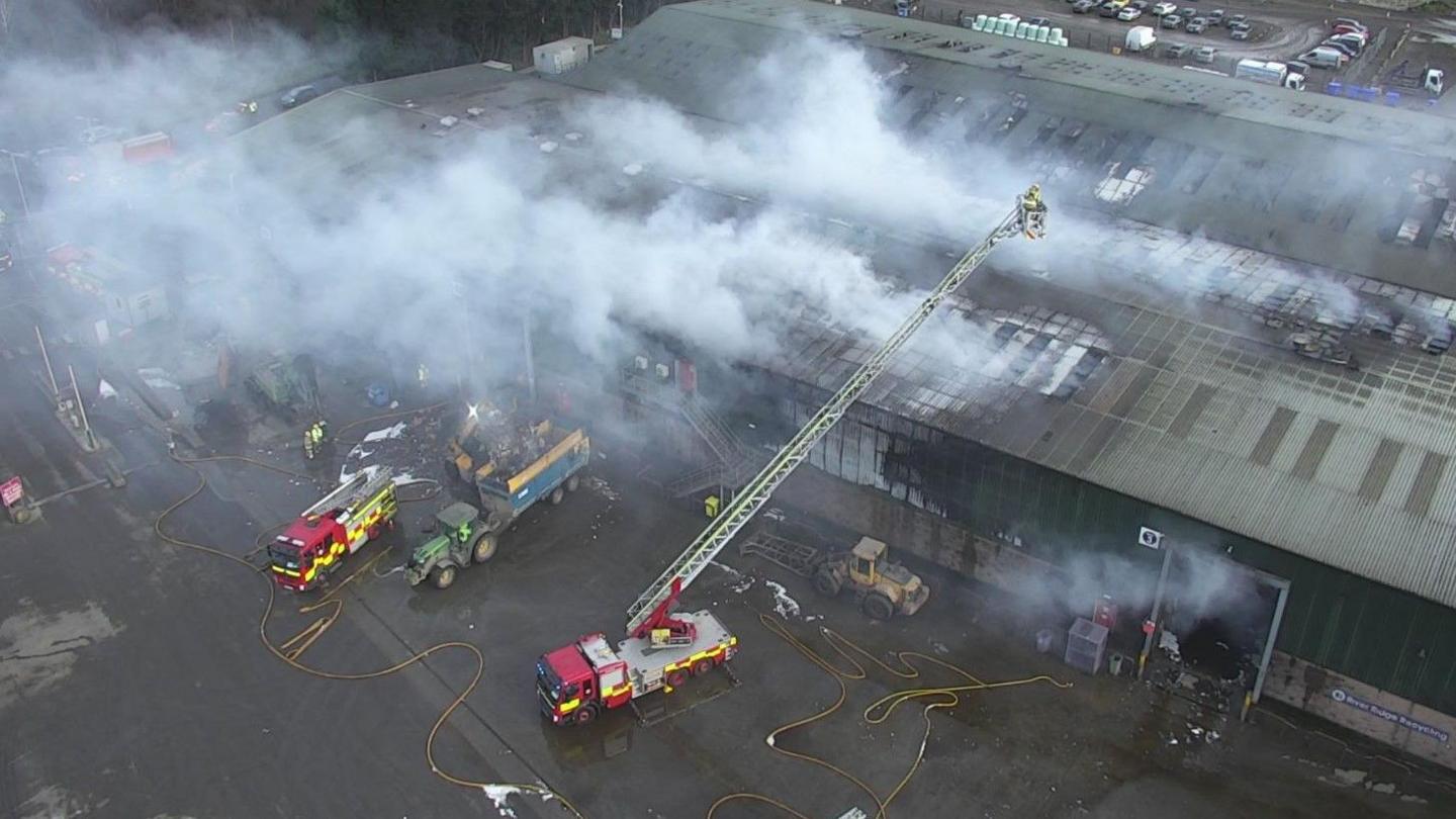 A number of fire appliances can be seen close to a smoking commercial premises. A fire engine's ladder is deployed over the building and a lone firefighter is on the ladder.