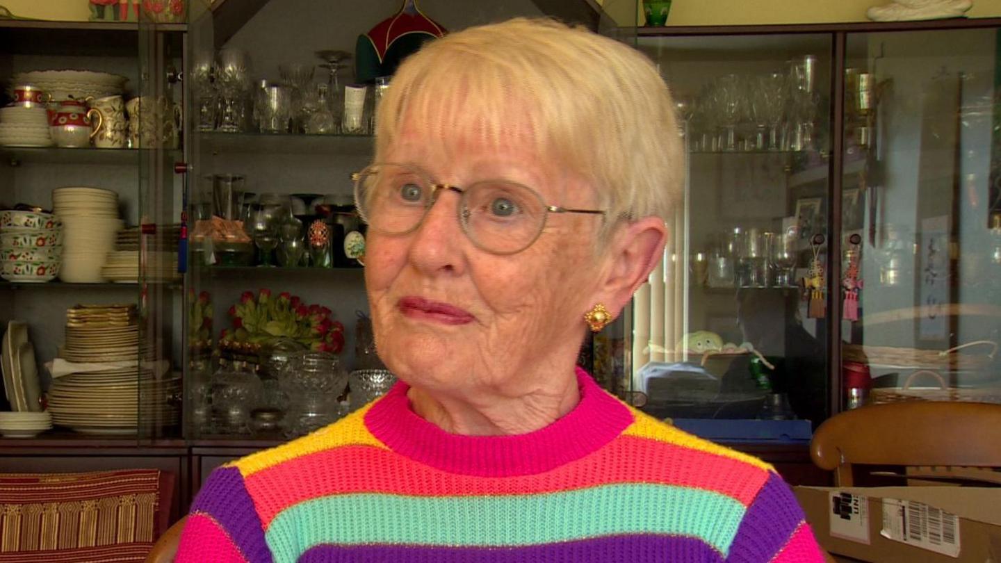 Christine Turner is an older woman with short blonde hair. She is wearing a pair of gold stud earrings and a pair of thin mental framed glasses. She is sitting in front of a large glass cabinet filled with glasses, plates and teapots/cups. She is wearing a bright pink, yellow, purple and turquoise striped jumper. She has a neutral expression on her face as she is turned to the side.